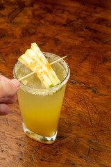 Close-up of a Hand Holding a Fresh Guava Cocktail in a Salt-Rimmed Tall Glass Garnished with Green Guava Sticks on Wooden Table