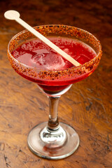 Close-up of a Spicy Red Cocktail with Ice and Wooden Stirrer in a Margarita Glass with Chili Powder Rim on a Rustic Wooden Table