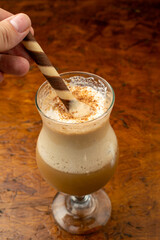 Hand stirring a creamy iced coffee frappe with a wafer stick, a cold dessert beverage with whipped...