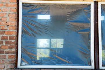 A window covered with plastic wrap shows reflections of sunlight and greenery, indicating neglect...