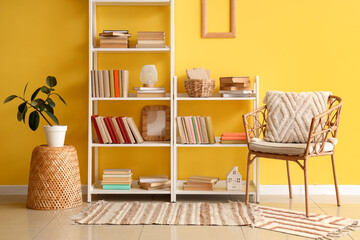 Shelving unit with books, armchair and houseplant near yellow wall in room