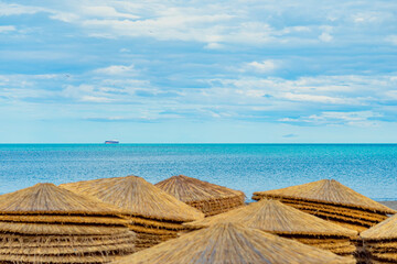 Traditional thatched beach umbrellas lined up on shore, cargo ship sailing on turquoise horizon....