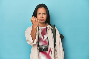 Filipina woman with camera and backpack on blue showing fist to camera, aggressive facial...