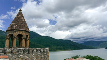 Ancient stone bell tower with arched windows overlooking a scenic lake and forested mountains under...