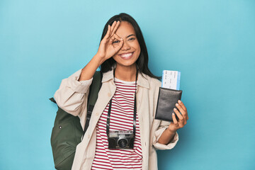 Filipina with camera, tickets, backpack on blue excited keeping ok gesture on eye.