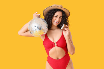 Beautiful woman in swimsuit, with coconut and disco ball on yellow background