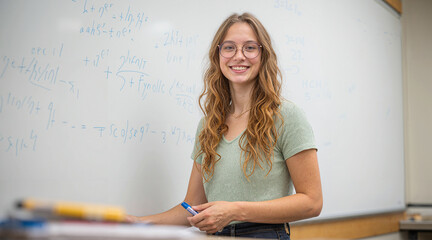A young woman stands near a whiteboard filled with complex equations, smiling confidently in a classroom or academic setting.