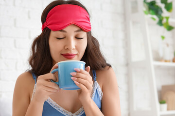 Beautiful young woman with sleep mask and cup of coffee in bedroom at home, closeup