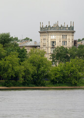 Classic architectural residential building with ornate decorations and sculptural rooftop details, partially hidden by trees along the river in Saint Petersburg.