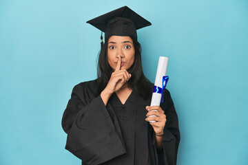 Filipina graduate with diploma on blue studio keeping a secret or asking for silence.