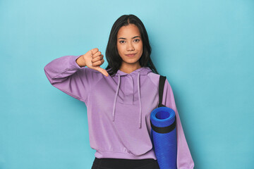 Filipina athlete with yoga mat on blue showing a dislike gesture, thumbs down. Disagreement concept.