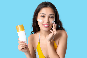 Beautiful young Asian woman in stylish yellow swimsuit applying sunscreen cream on her face against...
