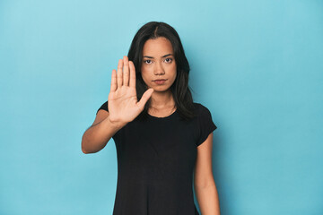 Filipina young woman on blue studio standing with outstretched hand showing stop sign, preventing...