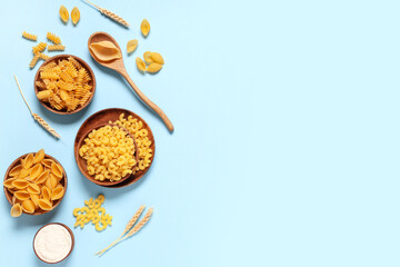 Wooden bowls with different types of raw tasty pasta, wooden spoon and spikelets on blue background