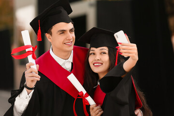 Couple of graduating students with diplomas taking selfie near university