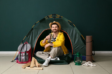 Young male tourist with tent, travelling equipment and thermos sitting on green background