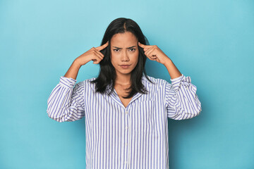 Filipina young woman on blue studio focused on a task, keeping forefingers pointing head.