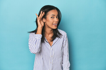 Filipina young woman on blue studio trying to listening a gossip.