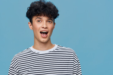 surprised young man with curly hair wearing a striped t-shirt against blue background expressing...