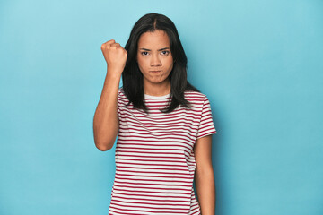 Filipina young woman on blue studio showing fist to camera, aggressive facial expression.