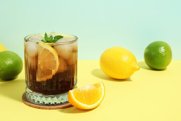 Glass of Long Island iced tea cocktail with mint and different citrus fruits on yellow table against turquoise background