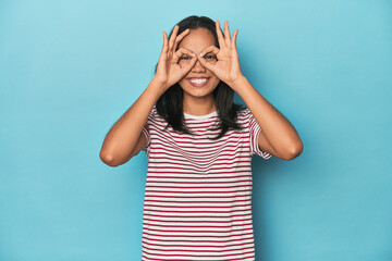 Filipina young woman on blue studio showing okay sign over eyes