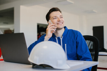 Smiling engineer talking on smartphone at desk with laptop and hardhat