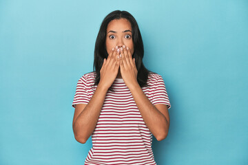 Filipina young woman on blue studio shocked, covering mouth with hands, anxious to discover...