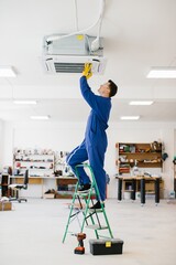 Electrician installing air conditioner unit on ladder in workshop
