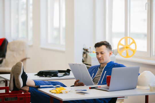 Worker relaxing with feet up, reading documents and using laptop in factory office