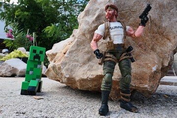 Fototapeta premium GI JOE action figure of survival instructor Outback, verion 2023 Classified edition, armed with pistol, ready to surprise explosive green LEGO Minecraft Creeper, while hidden behind a shoreline rock.