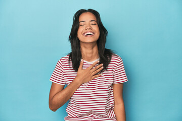 Filipina young woman on blue studio laughs out loudly keeping hand on chest.