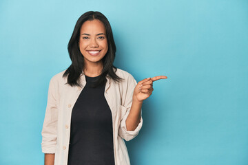 Filipina young woman on blue studio smiling cheerfully pointing with forefinger away.