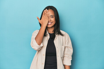 Filipina young woman on blue studio having fun covering half of face with palm.