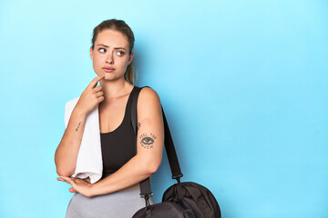 Sporty blonde with gym bag and towel in studio looking sideways with doubtful and skeptical...