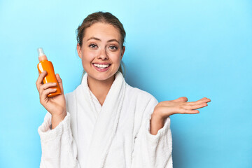 Blonde in bathrobe with sunscreen cream in studio showing a copy space on a palm and holding...