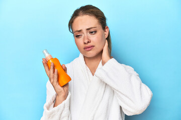 Blonde in bathrobe with sunscreen cream in studio touching back of head, thinking and making a...