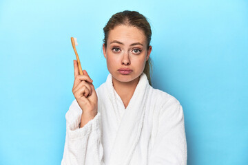Blonde in bathrobe with a toothbrush in blue studio shrugs shoulders and open eyes confused.
