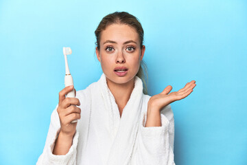 Blonde in bathrobe with electric toothbrush in studio surprised and shocked.
