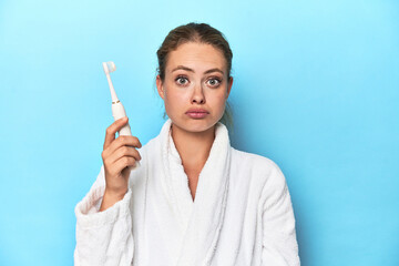 Blonde in bathrobe with electric toothbrush in studio shrugs shoulders and open eyes confused.