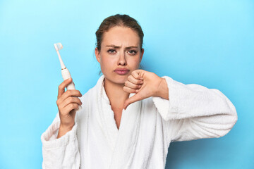 Blonde in bathrobe with electric toothbrush in studio showing a dislike gesture, thumbs down....