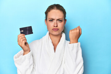 Blonde in bathrobe with credit card in blue studio showing fist to camera, aggressive facial...