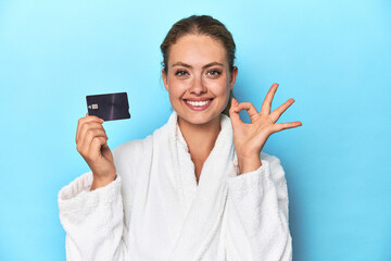 Blonde in bathrobe with credit card in blue studio cheerful and confident showing ok gesture.