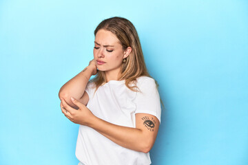 Blonde young woman in white t-shirt on blue background massaging elbow, suffering after a bad...