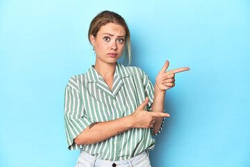 Blonde young woman in green striped shirt on blue background shocked pointing with index fingers to...