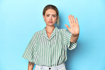 Blonde young woman in green striped shirt on blue background standing with outstretched hand...