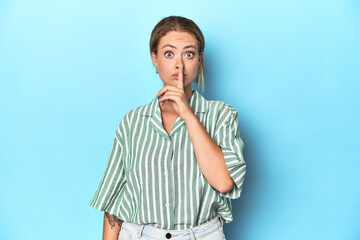 Blonde young woman in green striped shirt on blue background keeping a secret or asking for silence.