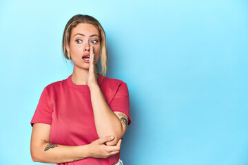Blonde young woman in red t-shirt on blue background is saying a secret hot braking news and...