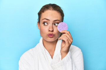Young blonde woman in a robe holding a facial sponge next to her face in a studio