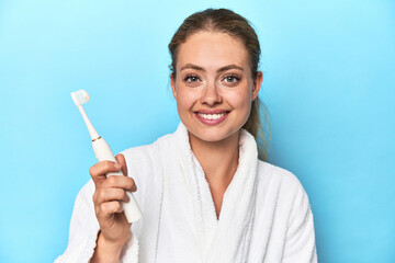 Young blonde woman in a robe holding an electric toothbrush in a studio
